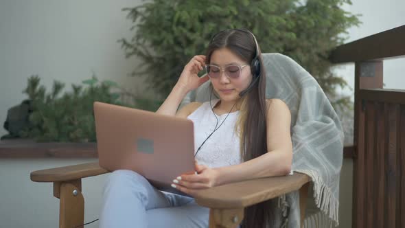 Charming Serious Asian Woman in Eyeglasses and Earphones Sitting on Armchair with Laptop Thinking