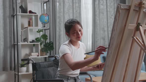 Side View Of An Asian Artist Girl In Wheelchair Holding Paintbrush Painting A Girl On The Canvas