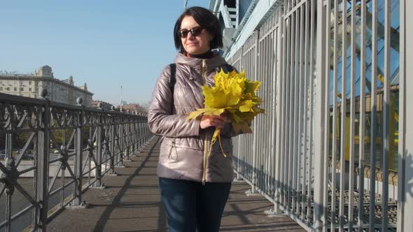 A Middleaged Brunette Woman in Black Glasses Walks Alone with a Bouquet of Yellow Leaves on the