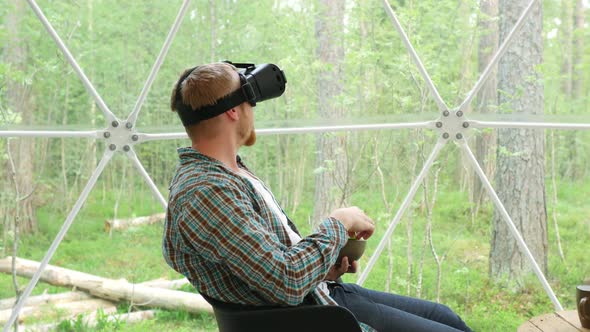 man in virtual reality glasses eats grapes in a dome tent overlooking the forest