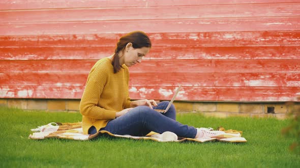 A girl with a laptop on her lap is sitting on a blanket on the lawn and typing on the keyboard