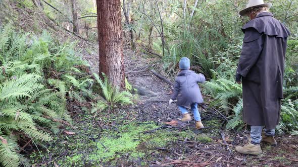 A bushman in a stockman jacket and an akubra hat jumps over a creek in a cold damp forest with his s
