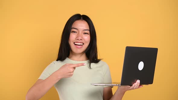 Amazed Happy Chinese Young Brunette Woman Holding an Open Laptop in Hand Pointing Finger at It Shows