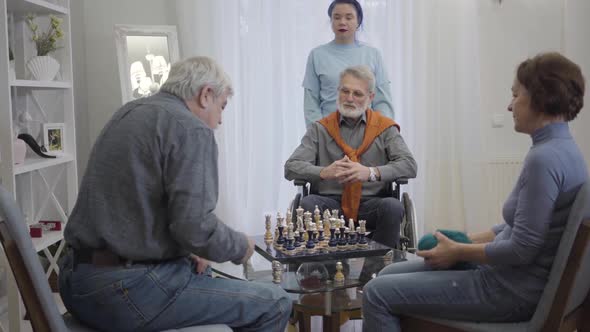 Young Female Volunteer Looking After Three Retirees in Nursing Home. Elderly Caucasian Men Playing