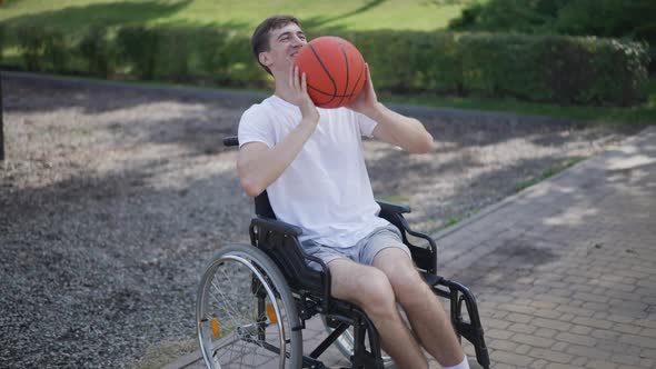 Portrait of Young Cheerful Caucasian Man in Wheelchair Catching Passing Basketball Ball in Slow