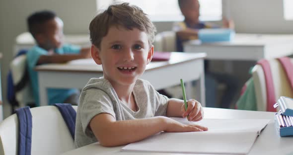 Video of happy caucasian boy sitting at desk during lesson in classroom