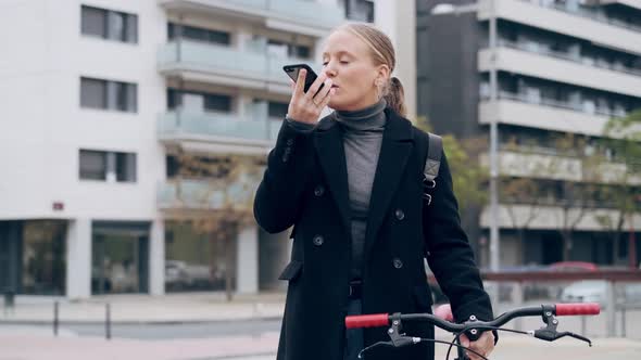 Woman pushing bicycle through city while speaking into smartphone