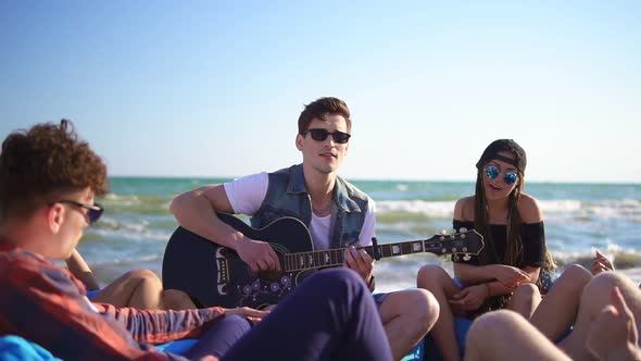 Young Man Playing Guitar Among Group of Friends Sitting on Easychairs on the Beach and Singing on a