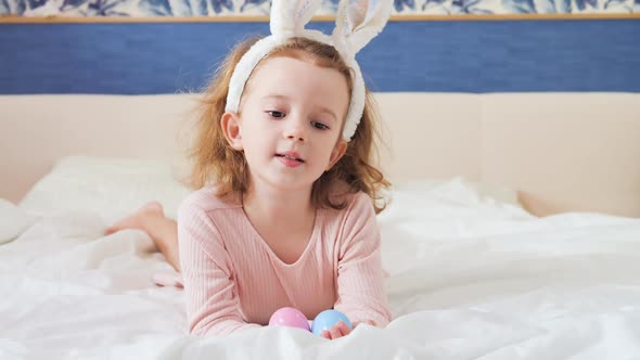 Cute Funny Girl Baby with Bunny Ears and Colorful Easter Eggs at Home on a White Bed