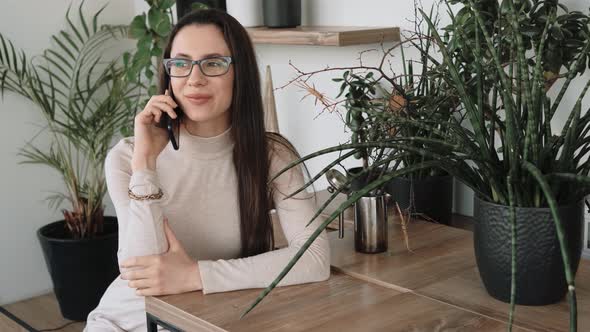 Young Woman Talking By Phone Sitting in the Green Office with a Lot of Plants on Background