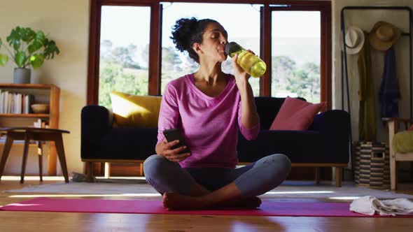 Mixed race woman drinking water and using smartphone while sitting on yoga mat at home