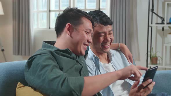 Asian Gentle Gay Couple Using Mobile Phone While Sitting On A Couch, Watching Funny Videos