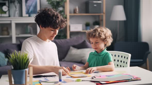 Young Woman Teaching Her Little Son To Make Paper Collage at Table at Home