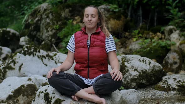 Calm woman in red vest meditates by the mountain river on vacation in nature. Mindful concept