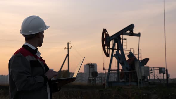 Man in a White Helmet with a Laptop on the Background of an Oil Rocking at Sunset