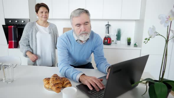 Portrait of Busy Senior Caucasian Man Messaging Online on Laptop As Loving Woman Hugging Husband