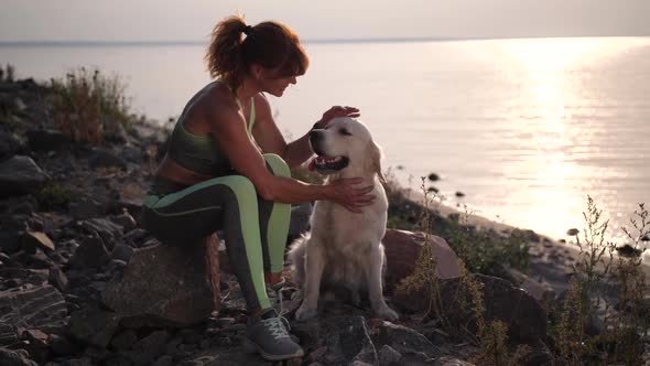 Fit Woman Stroking Dog Sitting on Shore at Sunset