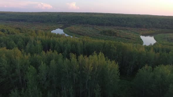 A drone flies low over a pristine forest and calm, winding river valley at sunset.