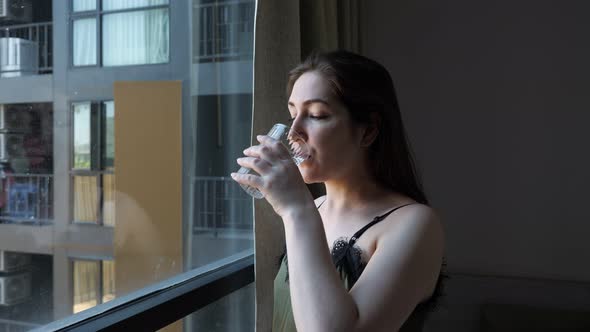 Happy Lady Drinks Water Looking Outside Window in Hotel Room