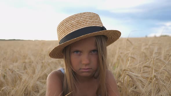 Offended Little Girl in Straw Hat Looking Into Camera Against the Background of Barley Field at