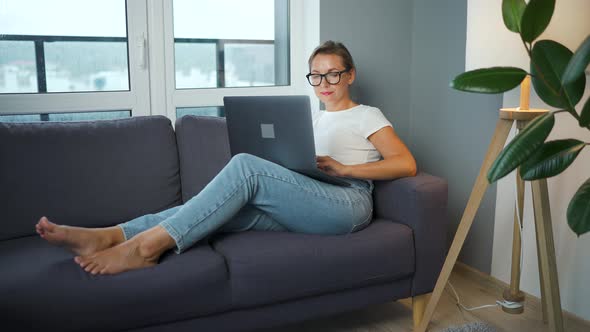 Woman with Glasses is Sitting on the Couch and Working on a Laptop