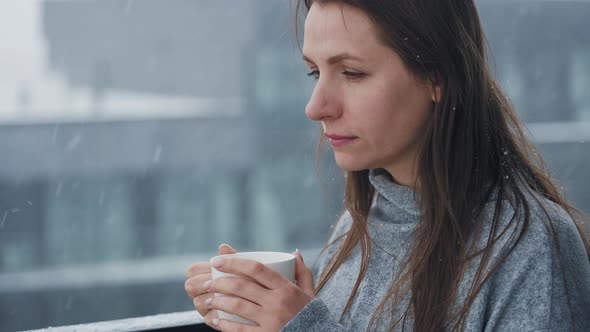 Caucasian Woman Stays on Balcony During Snowfall with Cup of Hot Coffee or Tea