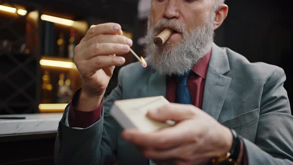 Senior Bearded Man Holding Burning Match and Setting Fire to Cuban Cigar Sitting at Own Cabinet Free