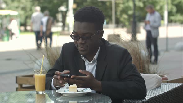 Handsome Young Afro American Businessman Using Smart Phone, Messaging His Girlfriend, Eating at Cafe