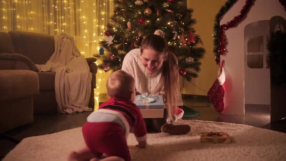 Little Baby Boy in Santa Claus Costume Crawling to His Mother Sitting Under Christmas Tree