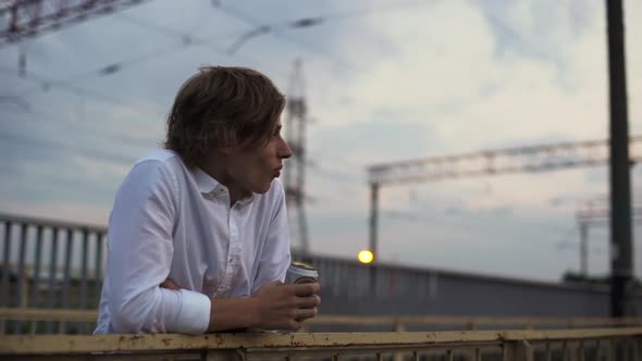 Blondhaired Handsome Man in White Shirt Standing on Bridge During Sunset and Drinking Beer From Can