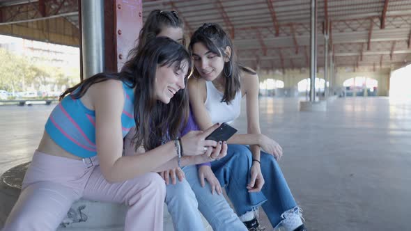 Three Happy Teenage Girls Sit and Look at Phone and Smile Outdoors