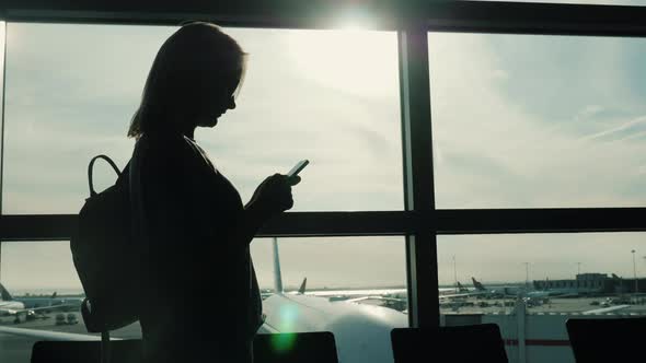 Always Online. Silhouette of a Woman Using a Smartphone in an Airport Terminal Near a Large Window