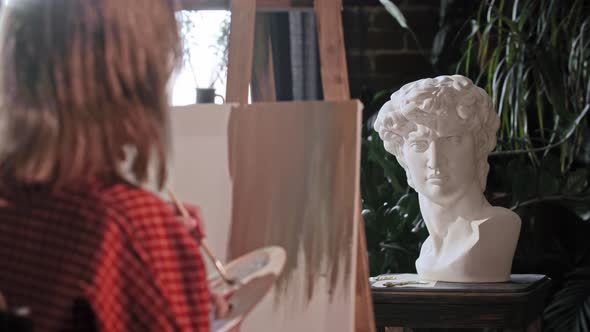 A Young Woman Artist Looking at a Greek Head Sculpture and Mixing Colors on a Palette