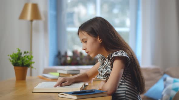 Side View of Pretty Schoolgirl Reading Book Doing Homework Sitting at Table at Home