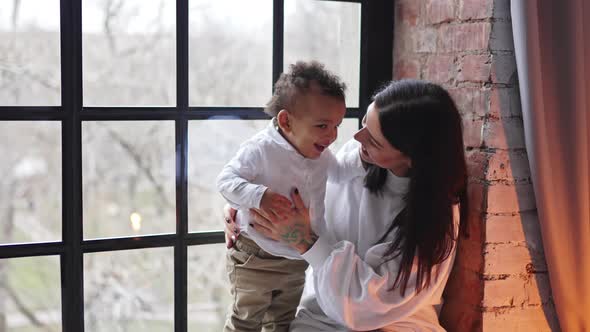 Satisfied Caucasian Mother Sitting on a Windowsill with Her Mixed Race Toddler