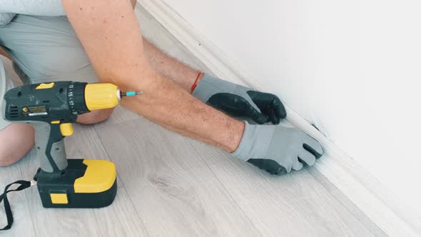 A Man Screws a Skirting Board to the Wall with an Electric Screwdriver in a Room Making Repairs in