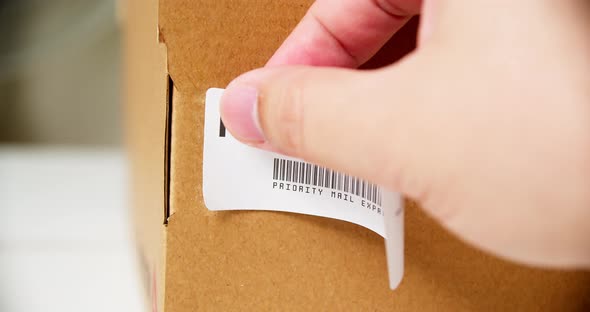 Hands applying PRIORITY MAIL Sticker label on a cardboard post box with barcode express.