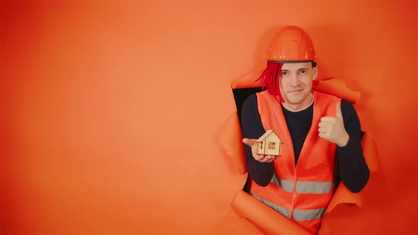 Male Construction Worker in Overalls Holding Small Wooden House and Sticking Out of Hole of Orange