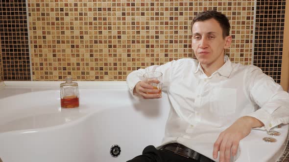 Drunk Man in Trousers and a Shirt Is Lying in the Bath and Drinking Alcohol