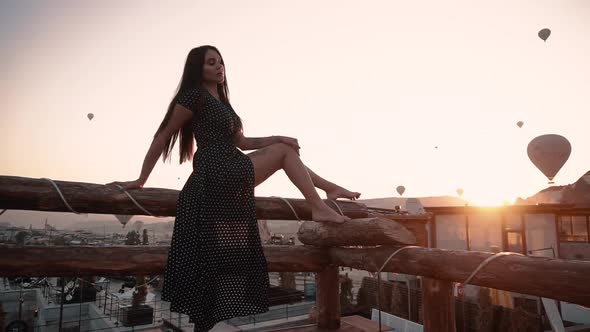 Beautiful Brunette with European Looks Sits on Terrace Background of Balloon at Sunset in Cappadocia