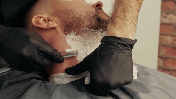 Close Up Barber Hands Shaving Bearded Man with Straight Razor in Male Salon