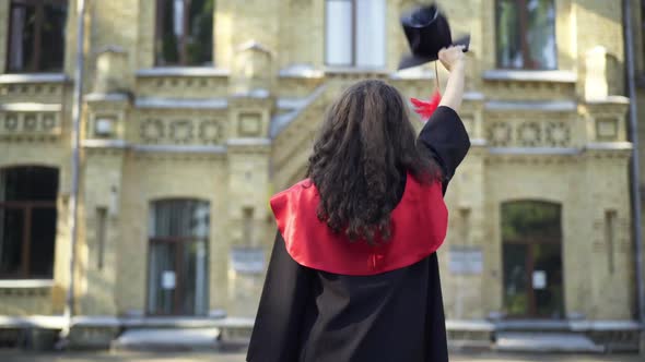 Back View Slim Young Woman in Graduation Toga Waving Goodbye with Mortarboard Cap to University