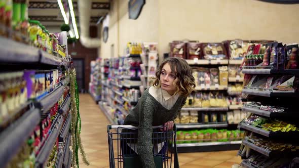 Front View of Caucasian Woman Walk with a Cart Near Shop Shelves Choosing a Glass Jar in Grocery