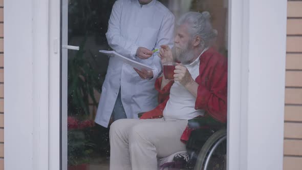 Senior Bearded Disabled Man in Wheelchair Drinking Tea As Nurse Bringing Documents for Sign