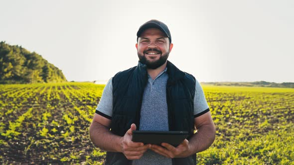 Happy Farmer Stands and Smile Holds Tablet in His Hands Against Background of Working Tractor in