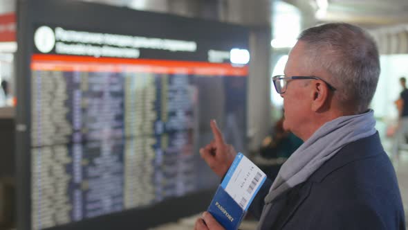 Senior Man with Ticket and Passport in Hand Looking at Information Board in International Airport