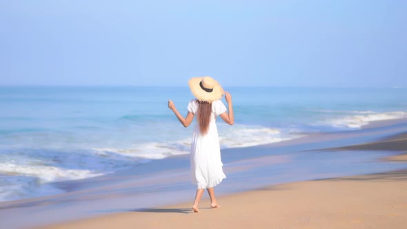 Back View Of Young Woman in White Sundress Walking Along the Sandy Beach by the Sea