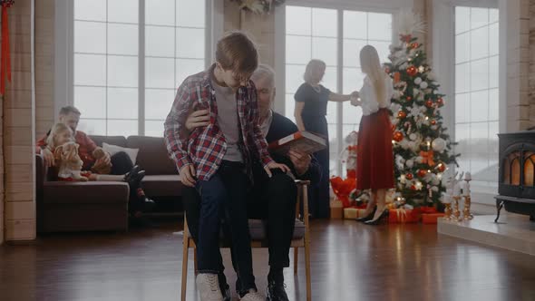 Boy Comes to Grandad Sitting on Chair to Read with Him at Christmas Celebration