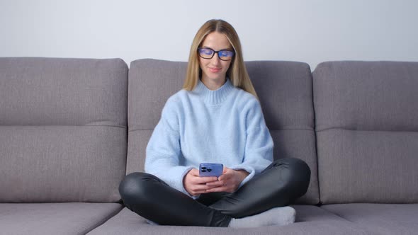 Happy woman using mobile phone while sitting on couch in apartment in 4k video