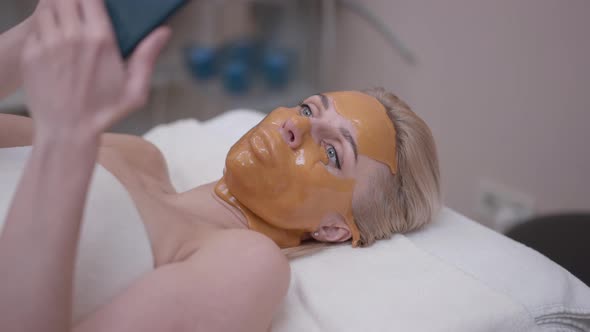 Beautiful Blond Woman with Dried Facial Mask Lying in Spa Salon Surfing Social Media on Smartphone